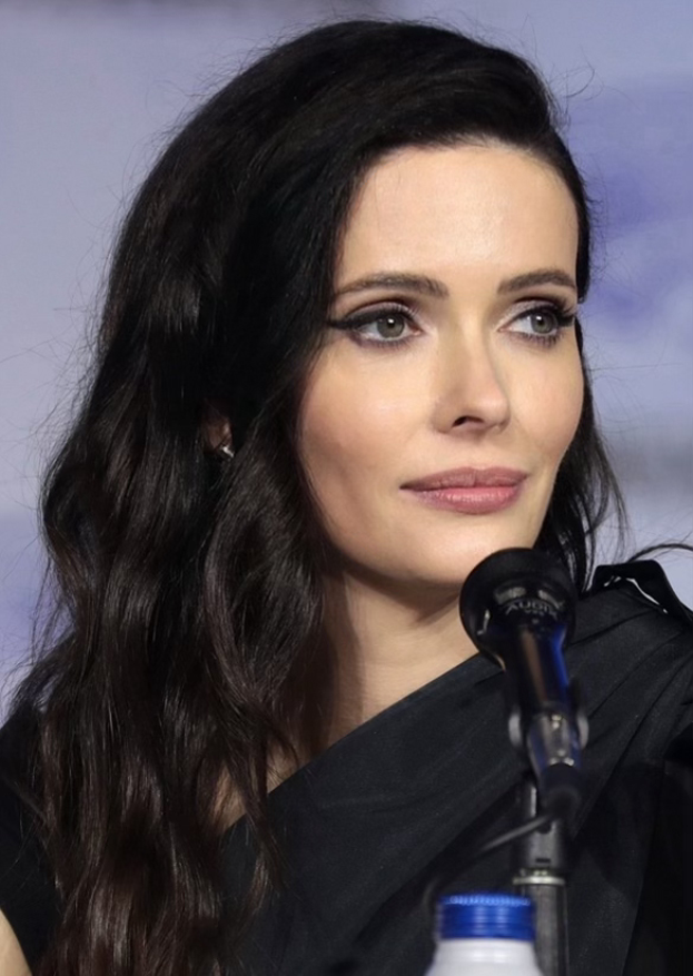 Krysten Ritter auf der Comic Con in San Diego, in einem schwarzen Kleid, vor einem Mikrofon mit einer Flasche unten, vor einem unscharfen, nebligen Hintergrund.