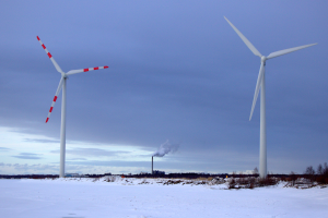 Drei Windkraftanlagen in einer verschneiten Landschaft, umgeben von Bäumen und Pflanzen, mit Rauch, der aus ihnen aufsteigt, unter einem bewölkten Himmel.