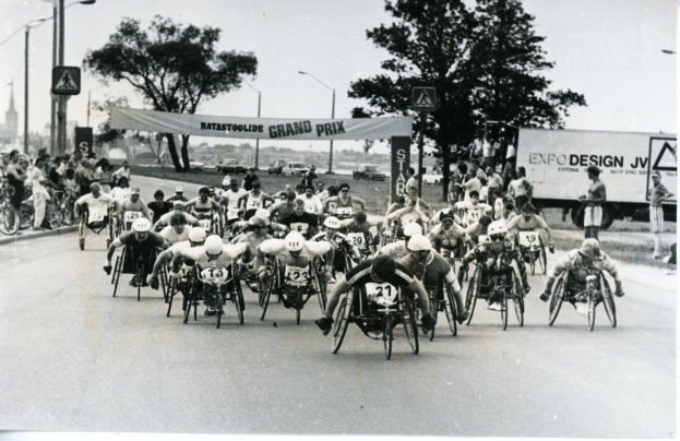 Eine Gruppe von Menschen im Rollstuhl fährt eine Straße entlang, trägt Helme und wird von einer Zuschauermenge am Straßenrand umgeben, auf einem Schwarz-Weiß-Bild.