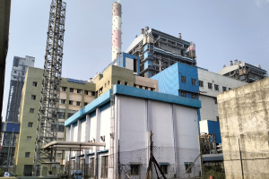 Ein großer Fabrikkomplex, wahrscheinlich ein kohlebetriebenes Kraftwerk, umgeben von Maschendrahtzaun, Masten und Bäumen, unter einem bewölktem Himmel, mit einem Wasserzeichen auf dem Bild, befindet sich in Indien.