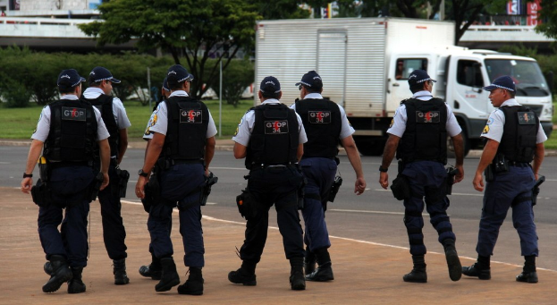 Eine Gruppe von Beamten in Uniform, darunter Mützen, Hemden, Schutzwesten, Uhren, Hosen und Schuhe, die auf einer Straße mit einem geparkten Lkw rechts, Gebäuden, Bäumen, Pflanzen, Masten, Bannern und einer Brücke im Hintergrund gehen.