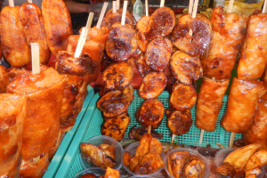 Marktstand mit Spießen voller Hühnerflügel und verschiedenen Lebensmitteln in Plastikbehältern, umgeben von Menschen und Flaschen.