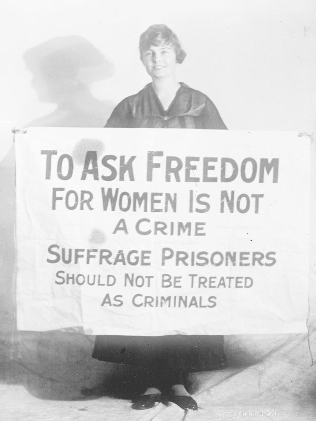 Schwarz-weißes Foto einer Frau, die ein Banner hält, auf dem "Für die Freiheit der Frauen zu kämpfen ist kein Verbrechen, Suffragetten sollten nicht wie Kriminelle behandelt werden" steht, während sie lächelt.