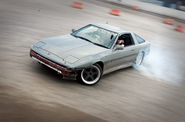 Eine Person in einem Helm fährt ein Auto auf einer Rennstrecke, gibt Rauch ab, umgeben von orangefarbenen Gegenständen, mit einem unscharfen Hintergrund, der hohe Geschwindigkeit andeutet.