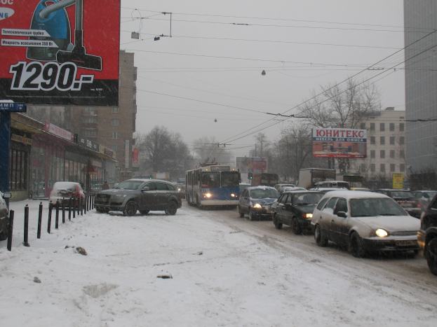 Eine von Schnee bedeckte Stadtstraße mit zahlreichen parkenden Autos, Gebäuden, Bäumen und anderen Objekten auf der linken Seite und mehr Gebäuden, Bäumen und Drähten im Hintergrund.