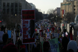Eine Gruppe von Menschen, die auf einer Straße protestieren und Plakate halten, mit Gebäuden, Bäumen, Laternen und dem Himmel im Hintergrund.