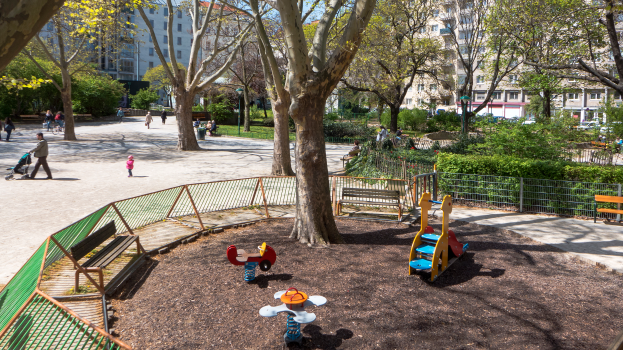 Ein Park mit einem Spielplatz in der Mitte, umgeben von Bäumen, Pflanzen, Geländern, Bänken und anderen Gegenständen, mit Menschen, einige halten Kinderwagen, und Gebäuden mit Fenstern im Hintergrund unter einem klaren blauen Himmel.