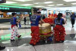 Eine Gruppe von Menschen in roter und blauer Kleidung geht durch einen Flughafen, einige tragen Musikinstrumente und Koffer, mit Geschäften, Namensschildern und Deckenlampen im Hintergrund, was auf eine chinesische Neujahrsfeier in Singapur hindeutet.