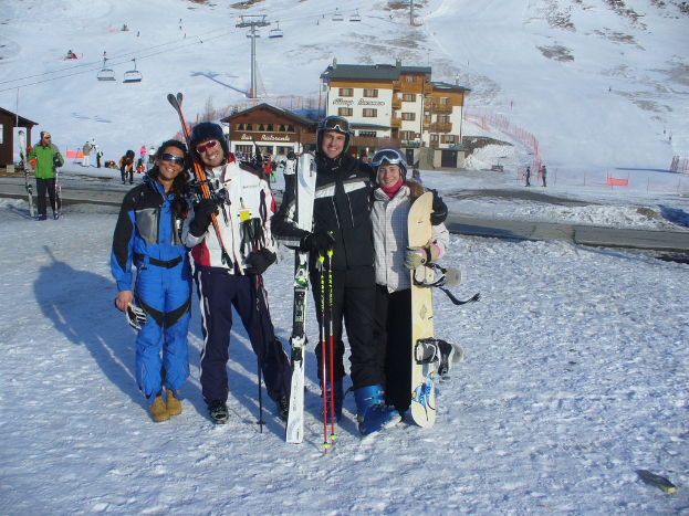 Vier Personen posieren mit Skaterausrütstücken und Werkzeugen für ein Foto, mit Gebäuden und anderen Menschen im Hintergrund, vor einem schneebedeckten Berg.