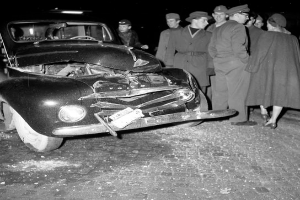 Ein Schwarz-Wei√ü-Bild eines verungl√ľckten Autos am Stra√üenrand, mit eingedellter Motorhaube und zersplitterten Fenstern, umgeben von einer Gruppe besorgter Menschen, die den Schaden begutachten.