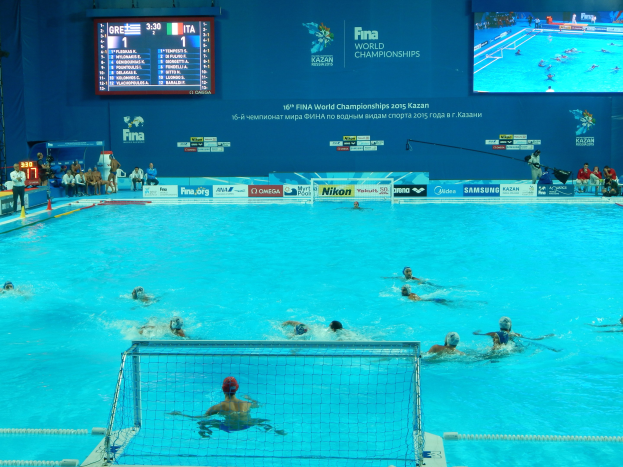 Eine Gruppe von Menschen spielt Wasserball in einem Pool während der FINA-Weltmeisterschaften 2016 in Kazan, mit Zuschauern auf der linken Seite und einer Anzeigetafel mit Event-Branding im Hintergrund.