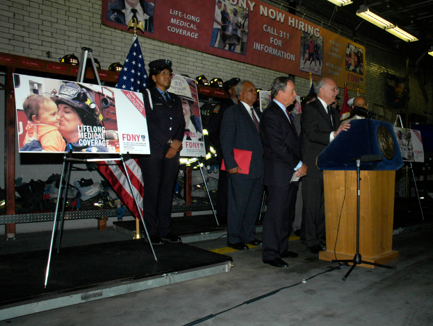 Eine Gruppe von Menschen steht in der Nähe eines Podiums mit Mikrofonen, umgeben von Schildern auf Ständern, Flaggen und Schildern an der Wand, alles unter hellen Scheinwerfern.