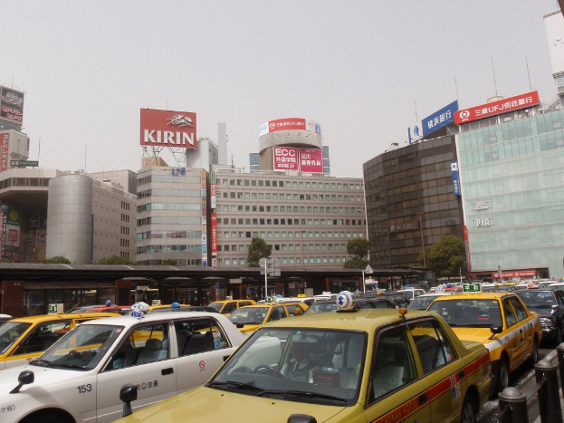 Eine Stadtansicht mit Fahrzeugen auf der Straße, Gebäuden, Bäumen, Polen mit Schildern und Text auf den Schildern, einem Zug im Hintergrund und einem Geländer rechts unten.