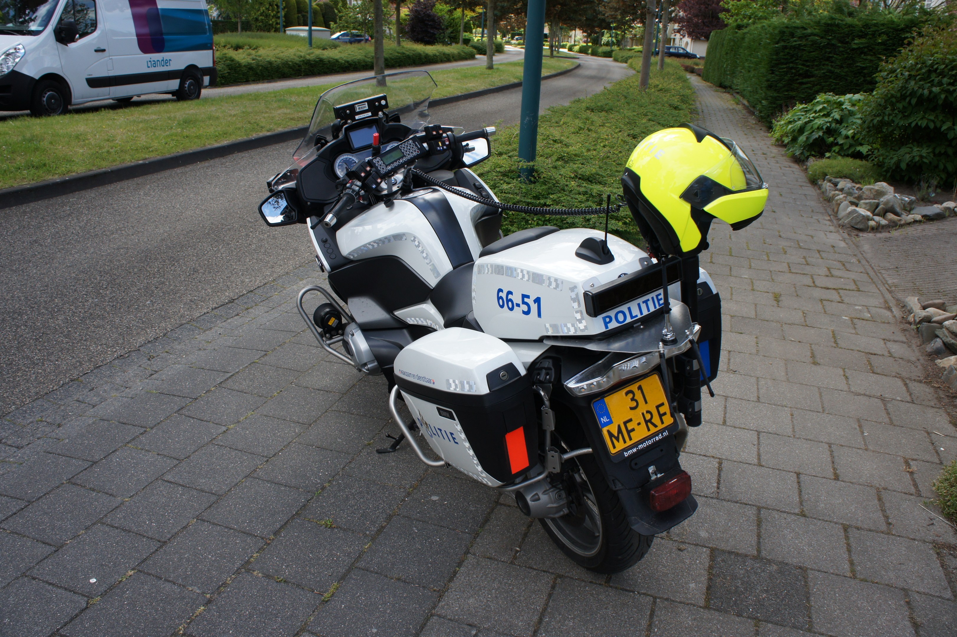 Eine Polizei-Motorrad auf dem Seitenstreifen geparkt mit einem Helm darauf, umgeben von Bäumen, Pfählen, Gebäuden, Pflanzen, Gras, Steinen und anderen Fahrzeugen im Hintergrund.