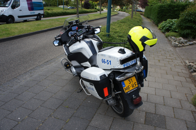 Eine Polizei-Motorrad auf dem Seitenstreifen geparkt mit einem Helm darauf, umgeben von Bäumen, Pfählen, Gebäuden, Pflanzen, Gras, Steinen und anderen Fahrzeugen im Hintergrund.
