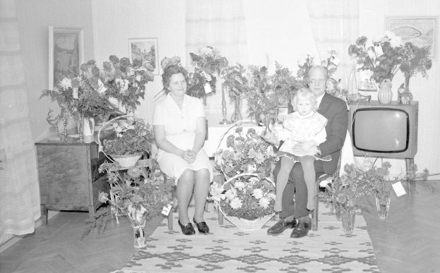 Ein Schwarz-Weiß-Foto einer Familie von drei Personen, die auf einem Sofa vor einem Fernseher sitzen, mit Blumenvasen auf nahen Tischen und gerahmten Bildern an der Wand.