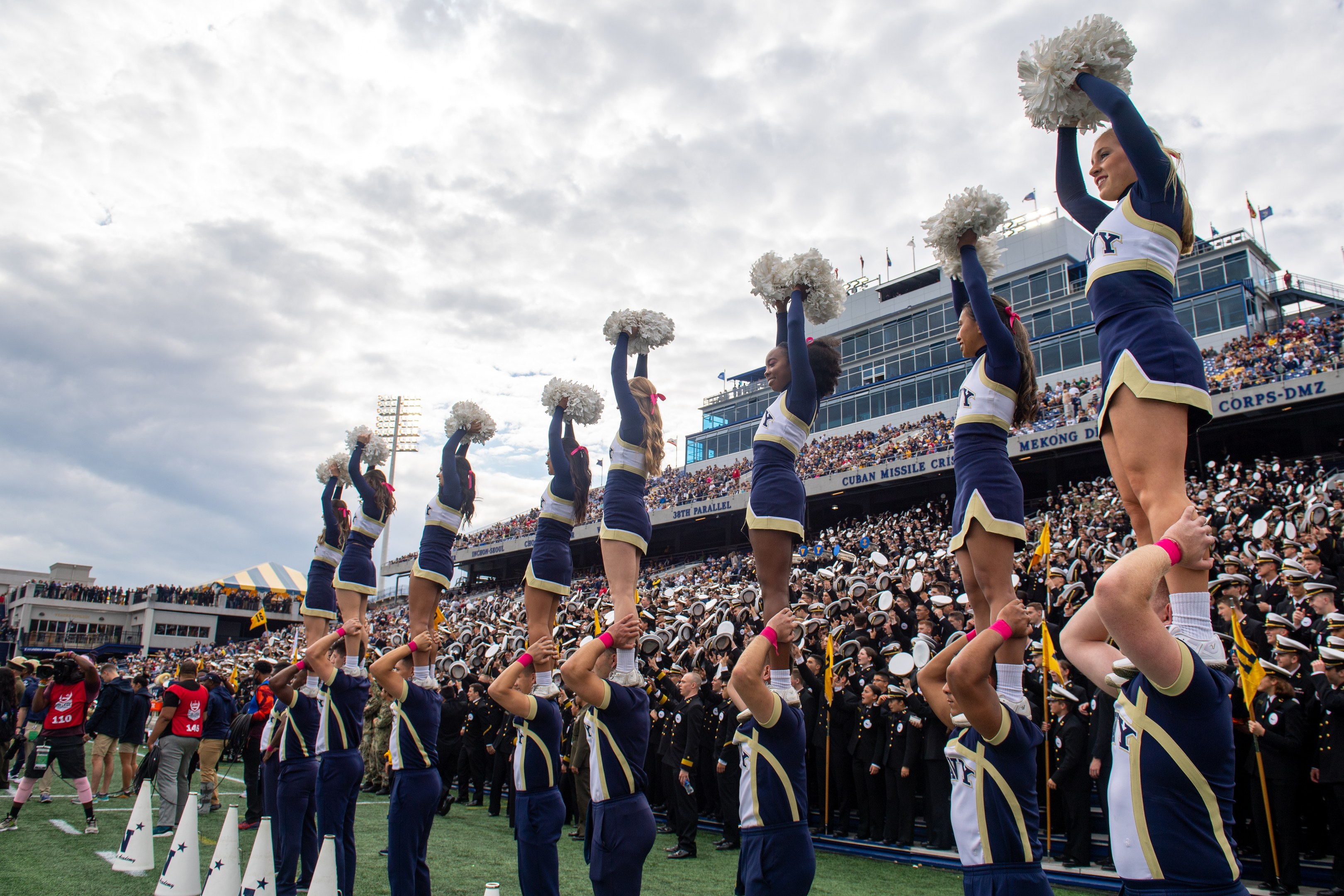 Eine Gruppe von Cheerleadern in blauen und weißen Uniformen führt einen Stunt auf einem Stadionevent durch, während sie Pompons halten und eine Menge zusieht und eine Person die Szene filmt.