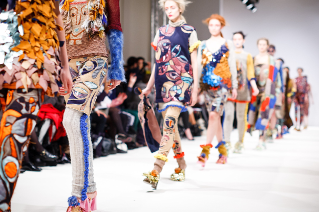 Eine Gruppe Von Frauen Geht Den Laufsteg Bei Der London Fashion Week entlang, Mit Einer Zuschauermenge Im Hintergrund Unter Helleren Lichtern.