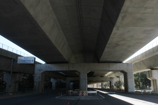 Autobahn unter einer Brücke mit Absperrgittern, Pollern und Texttafeln am Straßenrand, flankiert von Geländern, Bäumen und einem klaren blauen Himmel im Hintergrund.
