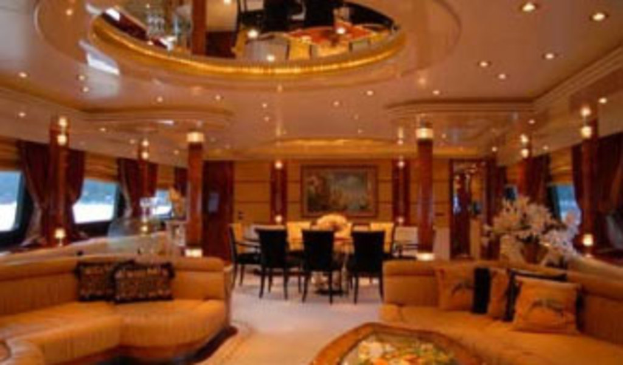 Ein Hotelfoyer mit Sofas, Stühlen, Tischen und Dekorationsgegenständen im Vordergrund, Fenstern mit Vorhängen und Lampen auf beiden Seiten, einem gerahmten Bild und Beleuchtung im Hintergrund sowie einem Spiegel mit Deckenbeleuchtung oben.