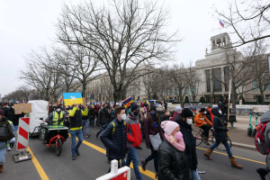 Eine große Gruppe von Menschen nimmt an einer Protestdemo auf einer Straße in Washington, D.C. teil und hält Schilder und Banner hoch, während einige Fahrräder fahren, Schilder, Bäume und ein klarer blauer Himmel im Hintergrund zu sehen sind.