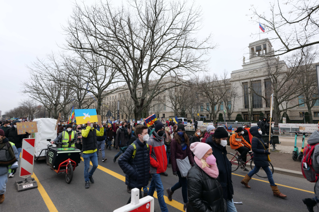 Eine große Gruppe von Menschen nimmt an einer Protestdemo auf einer Straße in Washington, D.C. teil und hält Schilder und Banner hoch, während einige Fahrräder fahren, Schilder, Bäume und ein klarer blauer Himmel im Hintergrund zu sehen sind.