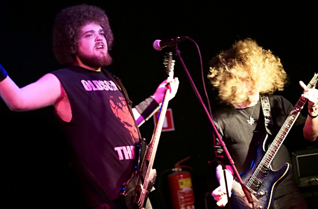 Zwei Männer, die auf der Bühne Gitarren spielen, beide in schwarzen T-Shirts, mit einem Mikrofon vor ihnen und einem Feuerlöscher im Hintergrund.