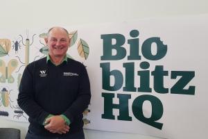 Ein Mann, der als Richard Geddes, CEO von Bio Blitz HQ, identifiziert wird, steht lächelnd vor einem Schild mit der Aufschrift "Bio Blitz HQ". Links von ihm befinden sich ein Stuhl und ein Tisch, und hinter ihm an der Wand befindet sich eine Tafel mit Text und Bildern.