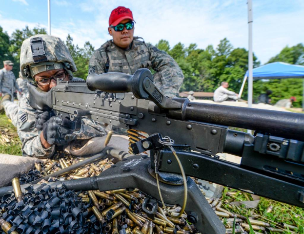 Eine Person auf einem Armee-Trainingsgelände feuert eine Waffe ab, mit einer anderen Person in einer roten Mütze daneben, während im Hintergrund Bäume, Zelte und Menschen zu sehen sind, unter einem klaren, sonnigen Himmel, mit Patronenhülsen und Gras im Vordergrund.