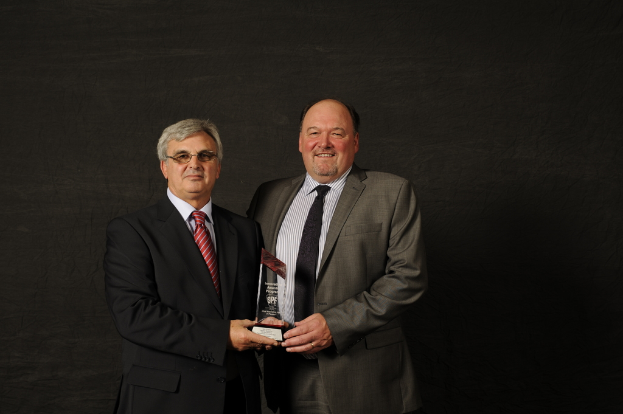 Zwei Personen in Anzügen und Krawatten, die stehen und einen Preis halten, mit einer Wand im Hintergrund.