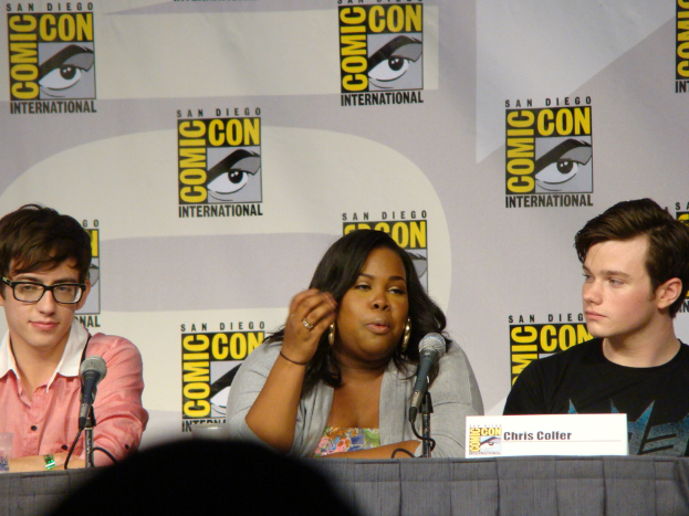 Eine Gruppe von Menschen sitzt an einem Tisch mit Mikrofonen, einem Namensschild und einer Werbetafel im Hintergrund, wobei ein Mann eine Brille trägt.