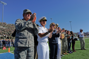 Eine Gruppe von Menschen auf einem Fußballfeld stehend, die klatschen, mit Zuschauern auf den Tribünen, Flutlichtmasten und einem klaren blauen Himmel im Hintergrund.
