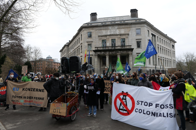 Große Gruppe von Menschen marschiert bei einer Demonstration gegen fossile Brennstoffe, trägt Schilder und Fahnen, mit einem Fahrzeug im Vordergrund und Gebäuden, Bäumen und einem klaren blauen Himmel im Hintergrund.