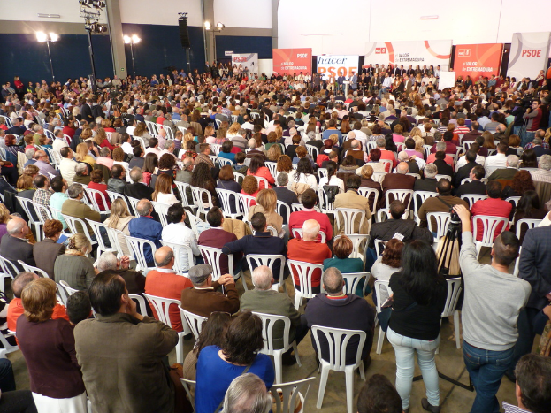 Eine Bühne mit Lichtern, einem Bildschirm und Musikern, die Instrumente spielen, mit einem Banner oben, einer Menge unten und Fahnen mit Stangen und Ballons drumherum.