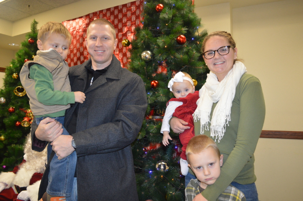 Ein Mann, eine Frau und Kinder stehen im Vordergrund mit Weihnachtsbäumen, Santa und einem Plakat im Hintergrund.