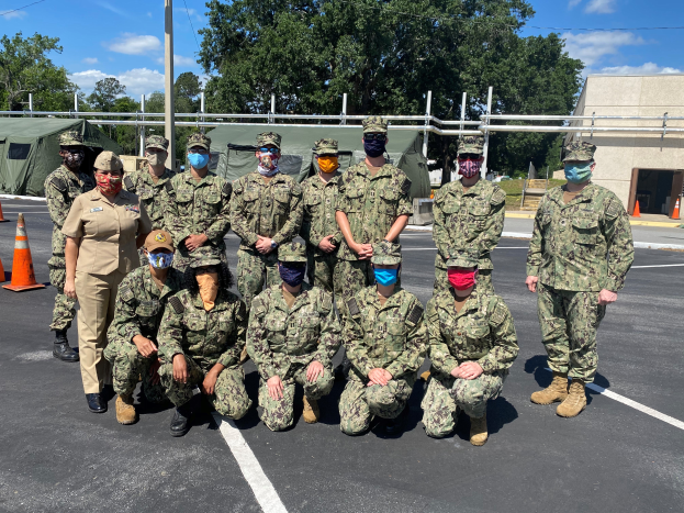 Eine Gruppe von Menschen in Militäruniformen, die Kappen und Masken tragen, posiert für ein Foto auf einem Parkplatz mit Verkehrskegeln links, Zelt, Pfosten, Bäumen, einem Gebäude und einem bewölkten Himmel, was auf ein Militärmanöver anspielt.