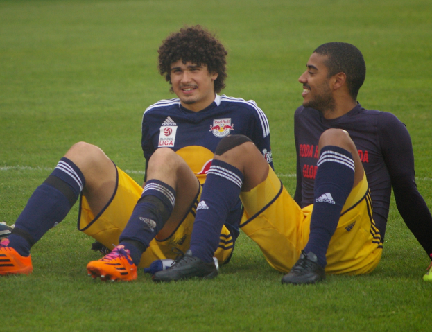 Zwei Fussballspieler sitzen im Schneidersitz auf dem Boden und tragen Sportkleidung und lacheln sich an.
