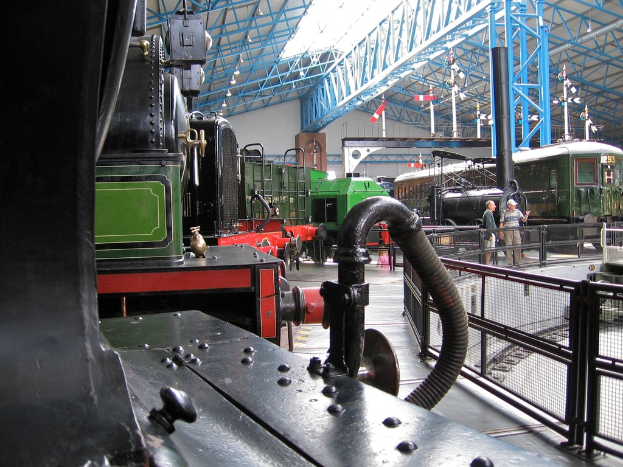 Mehrere Zugmaschinen sind in einem Abstellgleis geparkt, mit zahlreichen Eisenstangen oben. Rechts verläuft ein Zaun entlang einer Eisenbahnschiene mit einem Zug. Zwei Personen stehen in der Mitte der Szene nahe dem Zaun.