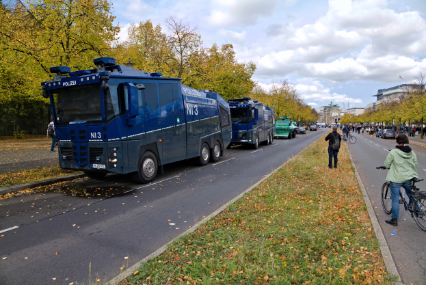 Eine Gruppe von Polizeiwagen, die auf der Seite einer Straße geparkt sind, mit einer Person, die auf einem Fahrrad auf der rechten Seite fährt. Es sind viele Menschen anwesend, einige halten Fahrräder, auf einem grasbewachsenen und blättertrockenen Boden. Im Hintergrund gibt es Bäume, Gebäude und einen bewölkten Himmel.
