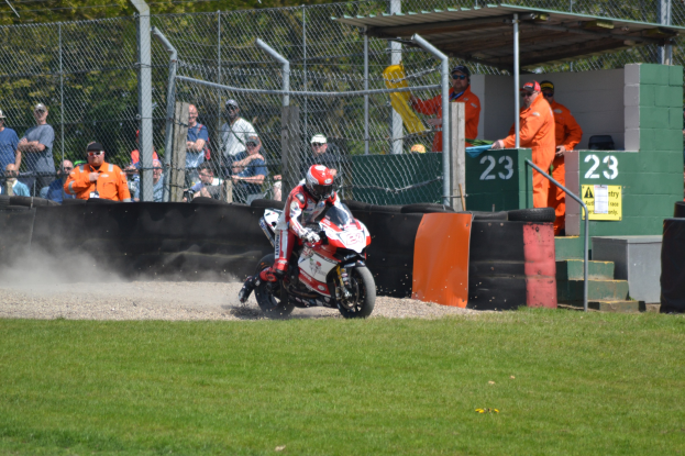 Eine Person in einem Helm und Handschuhen fährt ein Motorrad auf einem grasbewachsenen Feld, mit Zuschauern in Mützen und Schutzbrillen, einem Zaun, Treppen mit Geländern und einem nummerierten Schuppen mit Texttafeln im Hintergrund.