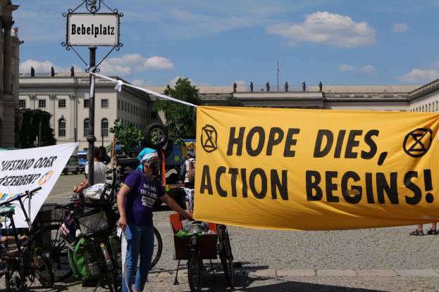 Eine Gruppe von Menschen steht vor einem Gebäude und hält ein gelbes Banner mit der Aufschrift "Hope Dies, Action Begins". In der Nähe stehen Fahrräder und ein Pfahl mit einer Tafel, vor einem klaren blauen Himmel mit Bäumen im Hintergrund.