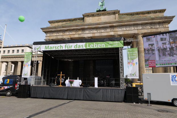 Bühne mit Tisch, Transparenten, Lautsprechern und Fahrzeugen vor dem Brandenburger Tor; Gebäude, eine Statue, ein Fahnenmast und ein bewölkter Himmel im Hintergrund.