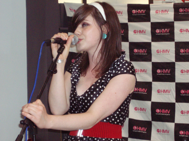 Eine Frau in einem schwarzen Kleid singt in ein Mikrofon, trägt ein Haarband und Ohrringe und hat ein Banner hinter sich.