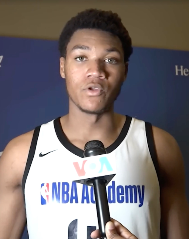 Ein Mann in einem weißen Tanktop hält ein Mikrofon und steht vor einer Banner, auf dem "NBA All-Star Game 2019" steht und einer Wand, wahrscheinlich spricht er mit der Medien.