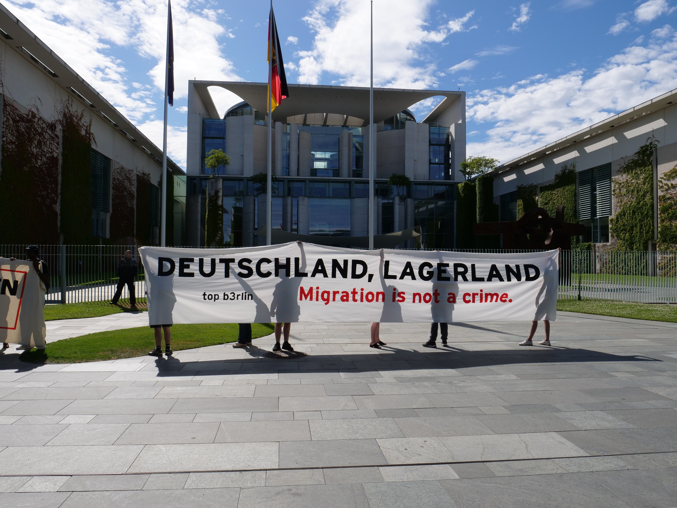 Gruppe von Menschen mit einem Banner, auf dem 'Deutschland, Lagerland Migration ist kein Verbrechen' steht, vor einem Gebäude mit Fenstern, umgeben von Gras, einem Metallzaun, Pflanzen, Bäumen und Fahnenmasten.