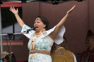 Eine Frau in einem weißen Kleid steht auf einer Bühne, schreiend in ein Mikrofon, mit einer Wand und einem Eisenrahmen im Hintergrund.