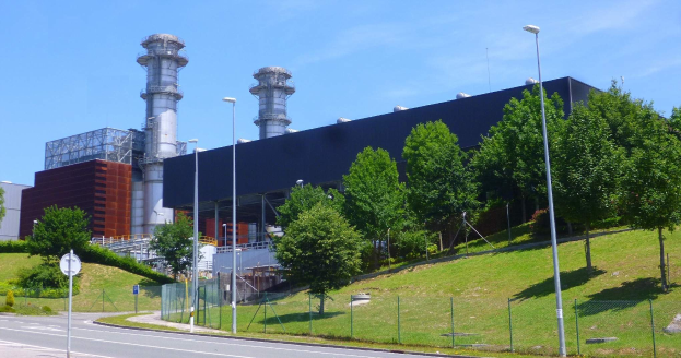 Großes Industriegebäude mit vielen Fenstern, als Kraftwerk identifiziert, umgeben von städtischer Infrastruktur einschließlich Straßenlaternen, Fahrzeugen und Grünflächen unter einem bewölkten Himmel.