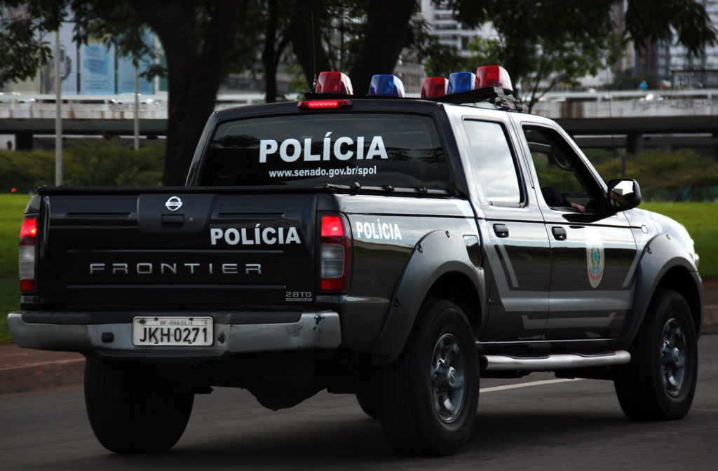 Ein schwarzes Auto fährt auf einer Straße mit sichtbaren Rädern, einem Spiegel, Gras, Bäumen, einer Brücke und Gebäuden im Hintergrund.