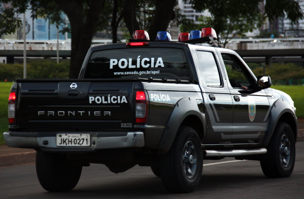 Ein schwarzes Auto fährt auf einer Straße mit sichtbaren Rädern, einem Spiegel, Gras, Bäumen, einer Brücke und Gebäuden im Hintergrund.