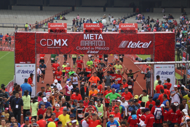 Eine große Gruppe von Läufern bei einem Marathon, mit einem Bogen in der Mitte, der einen Text anzeigt, einem Metallzaun auf der linken Seite und einigen Zuschauern auf der rechten Seite, mit leeren Stühlen im Hintergrund.